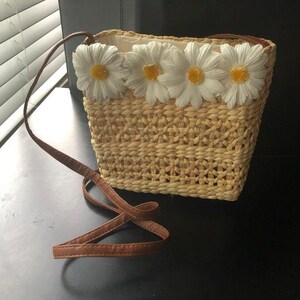 May include: A small, woven straw bag with a brown leather strap. The bag has a white fabric lining and is decorated with white daisy flowers.