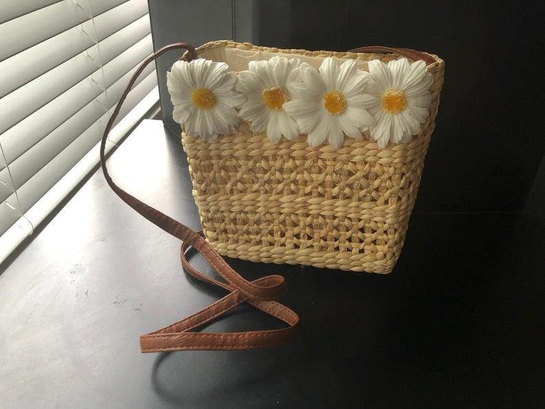 May include: A small, woven straw bag with a brown leather strap. The bag has a white fabric lining and is decorated with white daisy flowers.