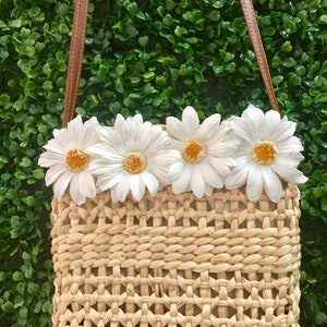 May include: A small woven straw bag with a brown leather strap and white daisy embellishments.