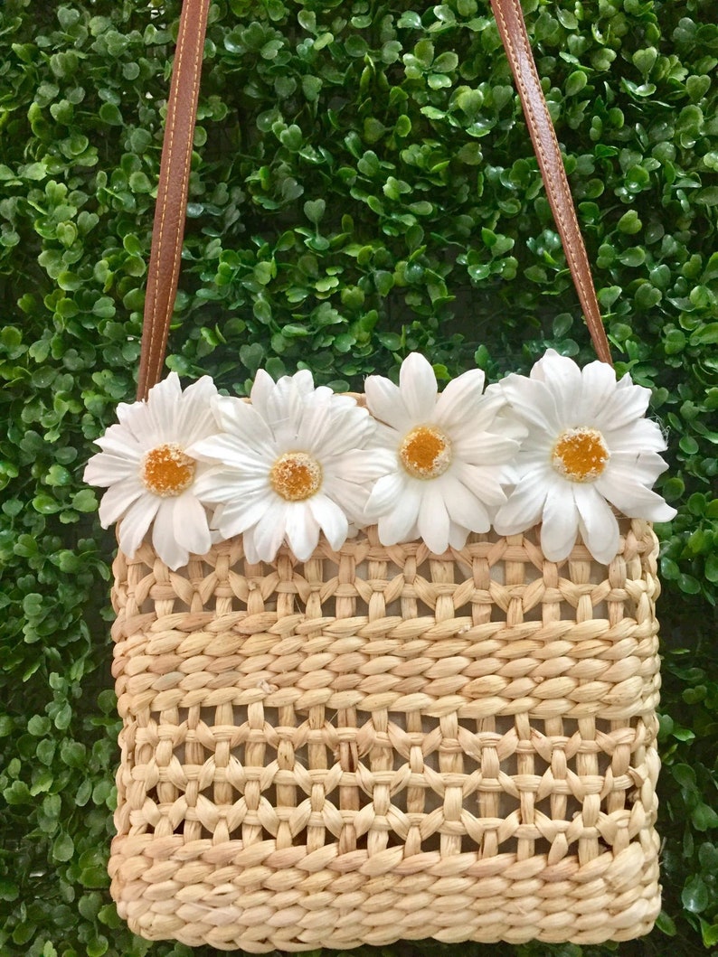 May include: A small woven straw bag with a brown leather strap and white daisy embellishments.