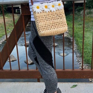 May include: A woven straw crossbody bag with a brown leather strap and a row of white daisy flowers across the front. The bag is being worn by a person wearing a striped shirt and grey leggings.