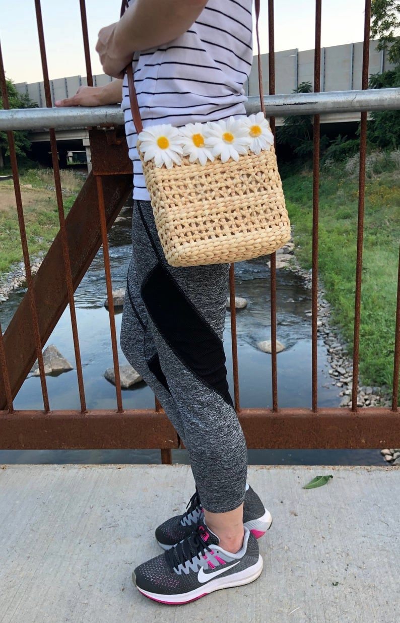 May include: A woven straw crossbody bag with a brown leather strap and a row of white daisy flowers across the front. The bag is being worn by a person wearing a striped shirt and grey leggings.