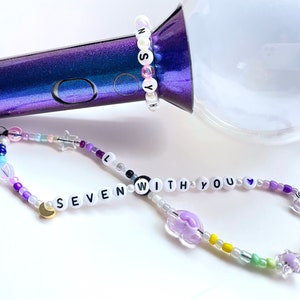May include: A beaded phone charm and bracelet set with the words "SEVEN WITH YOU" and "NSYH" spelled out in white letter beads. The charm features a variety of colorful beads, including stars, moons, and flowers. The bracelet is white with pink letter beads.