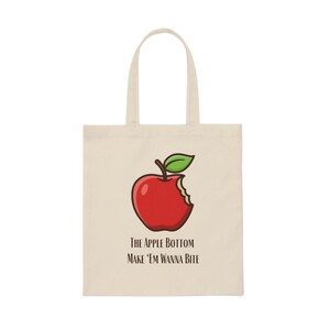 May include: A beige tote bag with a red apple with a bite taken out of it. The text on the bag reads "The Apple Bottom Make 'Em Wanna Bite".