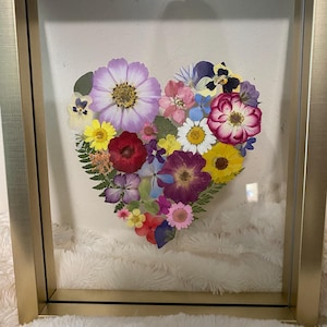 May include: A framed heart-shaped arrangement of pressed flowers in various colors, including purple, pink, yellow, and red. The flowers are arranged in a heart shape and are surrounded by a gold frame.