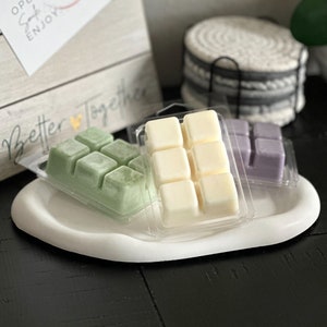 Puede incluir: Tres fundentes de cera en recipientes de plástico transparente. Los fundentes de cera son de color verde, blanco y morado. Los fundentes de cera están sobre una bandeja de cerámica blanca. El texto "Better Together" es visible en una caja de madera en el fondo.