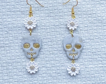 May include: A pair of dangle earrings featuring white polymer clay skulls with gold accents and white flower charms.