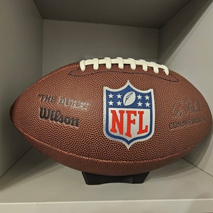 May include: A brown leather football with white laces and the NFL logo. The football is on a black stand and says "The Duke" and "Wilson" on the side.