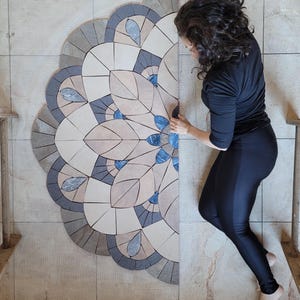 Może przedstawiać: Częściowo ukończony wzór mozaiki z płytek przedstawiający wzór kwiatowy lub mandalę. Projekt zawiera beżowe, niebieskie, szare i białe płytki. Mozaika jest instalowana na kafelkowanej powierzchni.