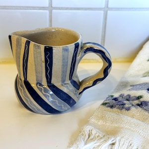 May include: A ceramic pitcher with a curved handle, painted with vertical stripes in shades of blue and cream. The pitcher has a unique, bulbous shape and a slightly irregular rim. A white towel with blue floral patterns is in the background.