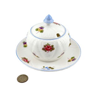 May include: A white ceramic sugar bowl with a lid and saucer, adorned with floral patterns in red, purple, and yellow. The bowl has a blue rim and a blue finial on the lid. A coin is in the foreground.