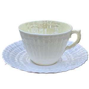 May include: A white teacup and saucer set. The teacup has a ribbed design and a curved handle. The saucer has a scalloped edge and a matching ribbed pattern. The set is made of porcelain.