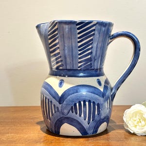 May include: A ceramic pitcher featuring a hand-painted blue and white pattern. The upper section displays vertical stripes, while the lower body is decorated with overlapping blue arches and dots. It includes a curved handle and a pouring spout.