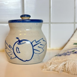 May include: A ceramic jar with a lid, featuring a hand-drawn apple design in blue. The jar has a cream-colored base with blue accents on the rim and lid. A fringed towel with floral embroidery is in the background.