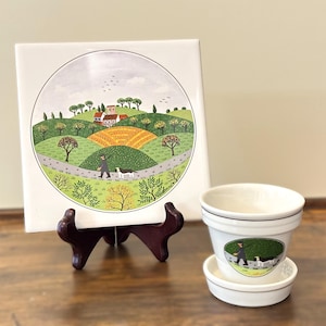 May include: A square ceramic tile featuring a circular landscape illustration of a person walking a dog on a path through rolling green hills, trees, and a small village, displayed on a dark stand next to a matching white ceramic flower pot.