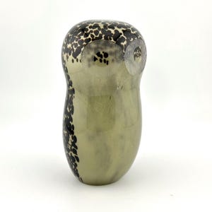 May include: A decorative owl-shaped glass figurine. The body is a pale green color with black speckled patterns on the top and along the side. The owl has a rounded shape and a smooth, glossy finish.