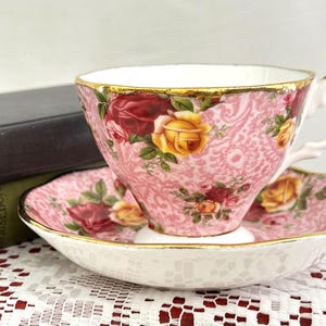 May include: A porcelain teacup and saucer set featuring a pink paisley-style background pattern with vibrant red and yellow rose floral motifs. The rim of both the cup and the saucer is finished with a thin, reflective gold-colored metallic trim.
