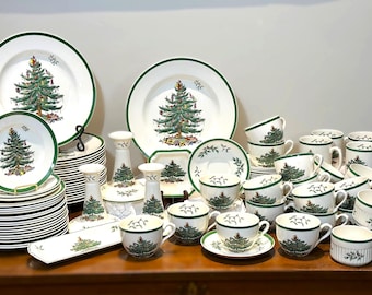 May include: A large collection of white ceramic dinnerware featuring a green rim and a central Christmas tree illustration. The set includes various sizes of plates, cups, saucers, bowls, and candlesticks, all decorated with matching festive motifs.