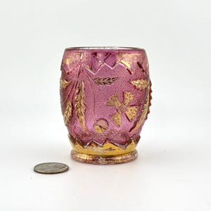 May include: A small, pink glass votive candle holder with gold leaf accents. The exterior features embossed floral and leaf patterns. The base and rim are also trimmed with gold. The candle holder is approximately 2.5 inches tall.