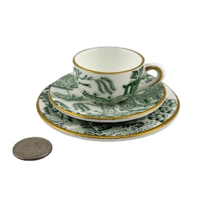 May include: A miniature tea set with a green and white Willow pattern. The set includes a teacup, a saucer, and a small plate, all with gold trim. The teacup is approximately 1 inch in diameter. The set is on a white background.