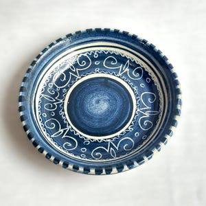 May include: A handmade ceramic bowl with a deep blue glaze and white decorative swirls. The rim features a textured, dark blue border. This decorative bowl is perfect for serving or display.
