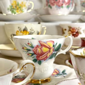 May include: A collection of white porcelain teacups and saucers featuring various floral patterns, including yellow and pink roses. Each cup has a gold-trimmed rim and base, arranged in a stacked display with multiple pieces visible in the frame.