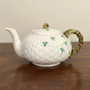 May include: A white ceramic teapot featuring a raised basket-weave texture, a green bamboo-style handle, and a matching spout. The body and lid are decorated with small, hand-painted green shamrock motifs and delicate gold-toned stems.