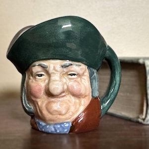 May include: A ceramic mug shaped like a smiling man's head, featuring a green hat, brown coat, and blue neckerchief. The mug has a handle and is set on a wooden surface. A book is visible in the background.