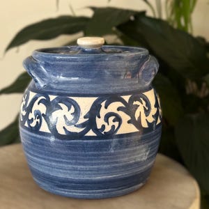 May include: A blue ceramic jar with a lid and handles. The jar features a white band with a blue scroll design. The lid has a small, round, off-white knob. The jar is sitting on a light-colored surface.