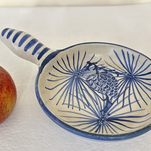 May include: A white ceramic serving dish with a blue and white pine cone design. The dish has a handle and is ideal for serving small portions of food.