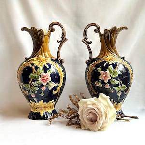 May include: Two antique ceramic vases with dark blue bodies, adorned with floral designs in pink, green, and gold. Each vase has a brown handle and a decorative top. A white rose and dried flowers are in the foreground.