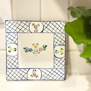 May include: Square ceramic plate with a white center and a blue and white lattice border. The center features a floral bouquet with yellow, pink, and blue flowers. Decorative plate for serving or display.