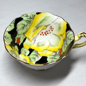 May include: A white teacup with a black, yellow, green, and red floral design. The cup has a gold rim and a gold handle.