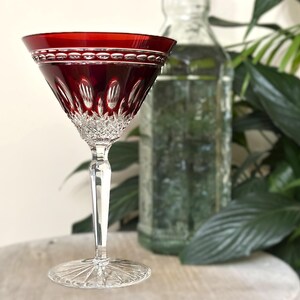 May include: A red crystal wine glass with a detailed pattern. The glass has a wide bowl and a long stem. The rim of the glass has a decorative border. A clear glass bottle is in the background.