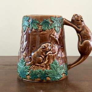 May include: A brown ceramic mug featuring a raised relief design of a squirrel on a tree trunk. The mug is decorated with green maple leaves around the top and bottom edges, and the handle is shaped like a squirrel climbing the side of the mug.