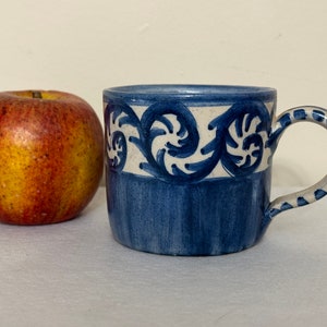May include: A blue and white ceramic mug with a decorative floral design. The mug has a handle and is approximately 9 cm tall.