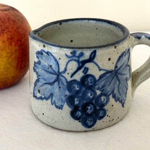 May include: A small, white ceramic mug with a blue and white grape design. The mug has a handle and is speckled with brown spots.