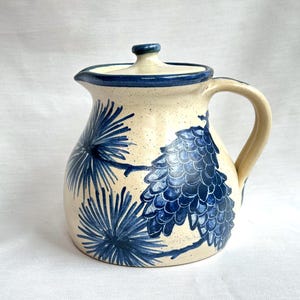 May include: Ceramic pitcher in cream with a lid, decorated with blue pine cone and branch motifs. The pitcher has a handle and spout, with blue detailing on the rim and lid. A decorative item for serving drinks, suitable for the kitchen.