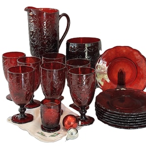 May include: A collection of ruby red pressed glass tableware featuring a pitcher, several stemmed goblets, a tumbler, and a stack of small plates. Each piece displays a raised floral pattern across the surface, creating a uniform look.