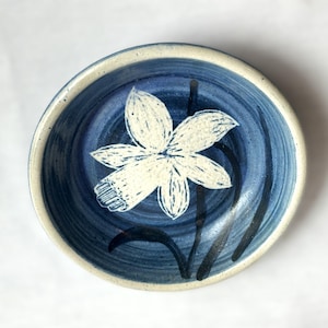 May include: A handmade ceramic bowl with a blue interior and a white floral design. The bowl has a light blue rim and a white flower in the center. The bowl is approximately 6 inches in diameter.
