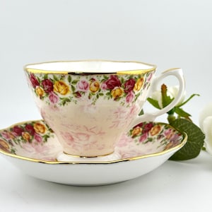 May include: A vintage teacup and saucer set with a floral rose pattern in pink, yellow, and burgundy. The teacup and saucer have gold trim. The set is on a white surface, with a white rose and greenery in the background.