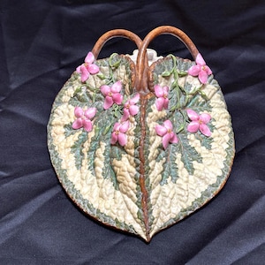 May include: A ceramic leaf-shaped tray with two brown handles. The tray has a detailed design of green and cream-coloured leaves with pink flower accents. The background is a dark, textured fabric.