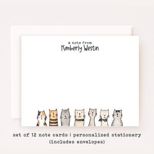 May include: A set of white note cards featuring the black printed text a note from Kimberly Westin at the top and a row of seven illustrated watercolor cats in various colors and patterns along the bottom edge, accompanied by matching envelopes.