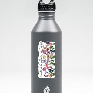 K&ouml;nnte beinhalten: Eine graue Mizu-Trinkflasche mit schwarzem Deckel. Ein Aufkleber auf der Flasche zeigt eine farbenfrohe Illustration von Wildblumen und den Text "Betrachtet die Lilien... Lukas 12,27."