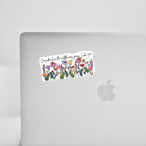 K&ouml;nnte beinhalten: Ein Aufkleber mit einer Aquarellillustration von bunten Wildblumen. Der Aufkleber hat den Text "Betrachtet die Lilien, wie sie wachsen. Lukas 12,27" in handschriftlicher Schrift. Der Aufkleber ist rechteckig mit wei&szlig;em Hintergrund.