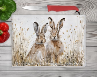 May include: A rectangular glass cutting board featuring a watercolor illustration of two brown hares standing in a field of tall, dry grasses. The artwork uses neutral earth tones with subtle paint splatters, depicting the hares facing forward.