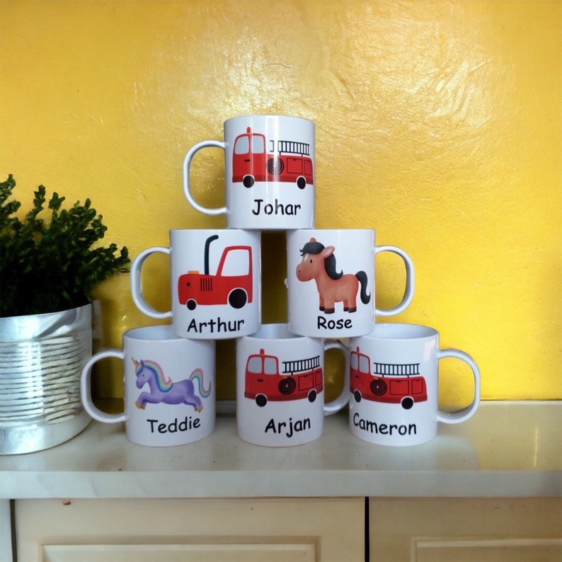 Op de afbeelding: Zes witte keramische mokken met kleurrijke ontwerpen en gepersonaliseerde namen. De mokken hebben een brandweerwagen, een eenhoorn, een tractor, een paard en een eenhoorn. De mokken zijn in een piramidevorm gerangschikt.