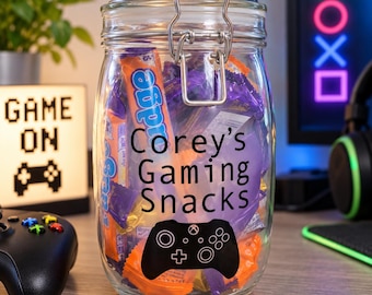 May include: A clear glass jar with a metal wire bail and lever closure, filled with assorted candy. The front of the jar features black text that reads "Corey's Gaming Snacks" above a black graphic of a video game controller silhouette.