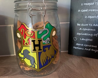 May include: A glass jar with a metal lid and a Hogwarts house crest sticker. The jar is filled with cookies. The sticker features the Hogwarts house crests for Gryffindor, Slytherin, Hufflepuff, and Ravenclaw. The jar is sitting on a wooden surface.