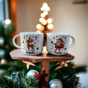 以下が含まれることがあります： クリスマスのデザインが施された白いセラミック製のマグカップが2つ。1つのマグカップには「Isabelle」という名前のトナカイが描かれ、もう1つのマグカップには「Charlie」という名前のサンタクロースが描かれています。